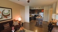 This single-floor residence offers a thoughtfully designed open-concept layout that seamlessly connects the living area and kitchen into one cohesive, inviting space. The living area features warm, earthy tones complemented by hardwood-style flooring that extends throughout, creating a sense of continuity and spaciousness. A ceiling fan overhead provides both function and a finishing touch to the room.

The kitchen is a standout feature, outfitted with dark espresso cabinetry that contrasts elegantly against lighter countertops and stainless steel appliances, including a refrigerator, microwave, and range. A peninsula with bar-height seating extends from the cabinetry, offering an ideal spot for casual dining or entertaining. Track lighting mounted to the ceiling illuminates the kitchen workspace effectively.

The open flow between the kitchen and living room enhances the overall sense of scale, making the space feel generous and well-connected. Neutral wall tones throughout contribute to a warm and welcoming ambiance, while the consistent flooring material ties the rooms together visually. The front entryway is conveniently located adjacent to the living area, offering easy access and a natural transition into the home's main living spaces.