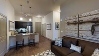 This single-floor residence features an open-concept layout that seamlessly connects the kitchen and living area, creating a cohesive and functional living environment. The space benefits from elevated ceilings that add a sense of airiness and volume throughout.

The kitchen is outfitted with dark cabinetry that contrasts sharply against light stone countertops, providing a modern aesthetic. A stainless steel refrigerator anchors the appliance suite, while a kitchen island with bar-height seating offers a casual dining option. Pendant lighting hangs above the island, adding both task illumination and a decorative touch. Track lighting further enhances the kitchen area.

The living area is anchored by a dark upholstered sofa dressed with patterned accent pillows, positioned atop a geometric area rug. A standout design element is the large reclaimed wood wall panel, which introduces rustic character and warmth to the otherwise contemporary space.

Warm-toned hardwood flooring flows consistently throughout, unifying the open floor plan. Neutral wall colors keep the space feeling bright and versatile. Multiple doorways visible along a central hallway suggest additional private rooms beyond the main living area.