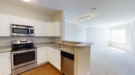 This single-floor, one-bedroom, one-bathroom residence offers a practical and well-designed layout centered around an open-concept living space. The kitchen is a standout feature, equipped with stainless steel appliances including an electric range, built-in microwave, and dishwasher. White cabinetry provides ample storage, while granite-style countertops add a polished finish. A double-basin sink is conveniently positioned along the counter, and a peninsula breakfast bar extends the workspace while creating a natural visual boundary between the kitchen and the adjoining living area. Track lighting overhead ensures the kitchen is well-lit. The transition from the kitchen's hardwood-style flooring to the plush carpeting of the living area creates a clear distinction between the two spaces. The open sightlines between the kitchen and living room contribute to an airy, connected feel throughout the main living area. Neutral wall tones and light-colored finishes throughout maintain a clean, move-in-ready aesthetic that complements a variety of décor styles.