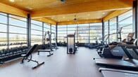 This well-appointed fitness center spans a generously sized single floor, thoughtfully divided into distinct functional zones. The facility's most striking architectural feature is its expansive floor-to-ceiling glass curtain walls, which flood the main workout area with abundant natural light and provide open views of the surrounding exterior landscape. Warm, natural wood-planked ceilings with recessed lighting and large ceiling fans complement the bright, airy atmosphere throughout.

The primary equipment area houses a full array of cardio machines, including treadmills, ellipticals, stair climbers, stationary bikes, and rowing machines, all positioned to take advantage of the panoramic glass walls. Strength-training equipment includes cable machines, a functional trainer, and free weights. A dedicated free weights and functional fitness zone features hardwood flooring and is stocked with dumbbells, medicine balls, resistance bands, stability equipment, and foldable mats.

A secondary, more open multipurpose area offers textured rubber flooring, light-toned stone-block accent walls, and a high vaulted wood ceiling with a statement oversized ceiling fan, providing ample open space for stretching or group exercise. Additional rooms off this area include private restrooms and storage, completing this comprehensive and well-designed fitness facility.