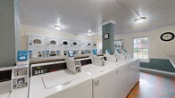 Located in Lyndon, Kentucky, this shared laundry facility is a single-floor space designed to efficiently serve a residential community. The room is lined with rows of commercial-grade top-load washing machines and stacked dryers, all featuring card-based payment systems for convenient, cashless use. The machines are arranged to maximize capacity while maintaining clear, accessible pathways throughout.

Along the perimeter, extended countertops provide generous folding and sorting surfaces, complemented by framed wall mirrors that add a sense of openness to the space. Seating is available along the walls, offering a comfortable waiting area for residents. A central structural column with a teal accent finish adds a subtle architectural detail to the otherwise utilitarian space.

The flooring features a distinctive multicolored diamond-pattern vinyl tile in shades of coral, teal, and green, lending a classic, retro character to the room. Natural light filters in through double-hung windows with views of surrounding greenery, brightening the interior alongside flush-mount ceiling light fixtures. Crown molding trims the walls, and a textured ceiling completes the tidy, well-maintained aesthetic. The overall layout is practical and functional, designed to accommodate multiple users simultaneously with ease.