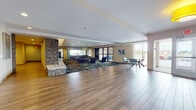 This single-floor common area is designed to serve as a central hub for residents, combining functionality with a contemporary aesthetic. The expansive lobby greets visitors with warm wood-look flooring, a striking stacked-stone accent column, and a teal-accented reception desk that adds a bold pop of color against neutral walls. Bold yellow drum pendant lights illuminate the space, contributing to a bright and inviting atmosphere.

The open lounge area transitions seamlessly from the hard-surface flooring onto a richly patterned carpet in deep teal, gold, and brown tones, anchoring a generous seating arrangement of dark leather sofas and modern accent chairs. Large floor-to-ceiling windows and glass sliding doors flood the lounge with natural light and offer views of the outdoors.

Adjacent to the lounge, a well-appointed kitchenette features dark cabinetry, stone-clad counters, a central island with a sink, and a row of stylish pendant lights overhead. A dedicated business center with workstations and a separate seating alcove with a stacked-stone column provide additional functionality. The mailroom includes a full wall of resident mailboxes alongside a package counter, rounding out this thoughtfully designed communal space.