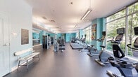Located at 10 Glover Ave in Norwalk, Connecticut, this fitness center offers a generously sized, single-floor exercise room designed to accommodate a full range of workout activities. The space is defined by its vibrant teal accent walls, durable rubber flooring, and an abundance of natural light streaming through floor-to-ceiling windows that line one side of the room, providing pleasant views of the surrounding greenery and streetscape.

The cardio zone is well-equipped with multiple treadmills, elliptical machines, and Matrix recumbent and upright bikes, all positioned to take advantage of the expansive window views. The strength-training area features a robust selection of Matrix selectorized machines, a cable crossover system, free weights with a dedicated dumbbell rack, adjustable benches, and mirrored walls that enhance the sense of space. Overhead, linear fluorescent light fixtures supplement the natural light to ensure consistent illumination throughout.

Colorful geometric artwork adds a dynamic visual element to the space, contributing to an energetic and motivating atmosphere. The layout is thoughtfully organized to separate cardio and strength zones, promoting efficient use of the floor area. A wall clock, whiteboard, and additional utility stations round out the functional amenities of this well-appointed fitness facility.