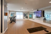Located in San Jose, CA, this well-appointed fitness center offers a thoughtfully designed workout environment across a single floor. The space is characterized by light hardwood flooring, bright white walls, and recessed lighting throughout, creating a clean and energizing atmosphere.

The main exercise area is open and spacious, providing ample room for a variety of workout activities. Stationary spin bikes are positioned for group or individual cycling sessions, while the open floor accommodates stretching and floor-based exercises. A large wall-mounted flat-screen television serves as a focal point on one wall, ideal for entertainment during workouts.

Full-length mirrors line key sections of the walls, enhancing the sense of openness and allowing users to monitor their form. A distinctive architectural column featuring bold navy and white vertical stripes adds a striking visual element to the space. A glass-railing mezzanine overlook connects to an adjacent lower-level fitness floor equipped with treadmills and additional cardio machines, visible below.

Natural light filters in through multiple windows, while the vibrant teal accent walls below add a pop of color to the overall palette. The staircase connecting the levels is refined, with metal railings and a large landscape-inspired mural artwork adorning the landing wall, providing an artistic transition between floors.