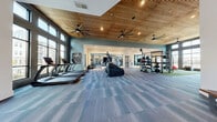 Located at 2370 Sever Rd in Lawrenceville, GA, this fitness center is a thoughtfully designed, single-floor facility offering a comprehensive workout experience across a generously sized floor plan. The space is divided into two distinct functional zones, each tailored to a specific style of training.

The cardio and general fitness area features an impressive architectural ceiling clad in warm wood slats, accented by large, statement ceiling fans and recessed lighting. Floor-to-ceiling windows flood the space with abundant natural light and offer views of the surrounding outdoors, creating an open and energizing atmosphere. Patterned carpet tile flooring runs throughout this zone, lending a clean, modern aesthetic. A row of treadmills, stair climbers, and other cardio machines from the True fitness brand are well-positioned for comfortable use.

The strength and functional training zone transitions to turf flooring, marked with agility lines for sport and performance training. This area is equipped with free weights, adjustable benches, cable machines, TRX suspension systems, stability balls, and a multi-functional rack system. A large mirror wall visually expands the space, while motivational phrases such as "Walk In Strong" and "Stronger" are prominently displayed on the walls. A wall-mounted television and a posted fitness center rules board add practical amenities. The high ceilings continue into this zone, maintaining the spacious and uplifting ambiance throughout the entire facility.