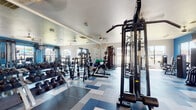 This single-floor fitness center is thoughtfully designed to accommodate a wide range of workout styles and fitness goals. The open layout provides ample floor space, allowing for the comfortable arrangement of both strength training and cardiovascular equipment without congestion. The flooring features a distinctive geometric pattern in shades of blue and gray, adding a modern, energetic aesthetic throughout the space.

The strength training area is well-stocked with a full rack of dumbbells, multiple Nautilus cable machines, weight benches, barbells, and plate-loaded equipment. Floor-to-ceiling mirrors line portions of the walls, enhancing the sense of openness and allowing users to monitor their form during workouts. Kettlebells and stability balls are also available for functional training.

The cardio section is positioned near large windows that invite natural light and offer views of the surrounding outdoor area. Treadmills, elliptical trainers, and a recumbent bike are arranged facing a wall-mounted television, making cardio sessions more engaging. Bold blue accent walls add a vibrant, motivating atmosphere to this zone.

Ceiling fans are distributed throughout the space to maintain airflow and comfort. A water fountain and additional amenities are accessible near the entrance, completing this fully functional and visually dynamic fitness facility.