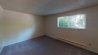 This single-floor residence at 32122 46th Pl SW in Federal Way, Washington, offers a straightforward and functional layout across several distinct areas. The living space features wall-to-wall carpet in a neutral brown tone, complemented by textured ceilings and baseboard heating units along the walls. A partial wall divides the main living area from a tiled hallway that leads to additional rooms, providing a degree of separation without fully enclosing the space.

The kitchen is efficiently arranged, featuring warm-toned wooden cabinetry, dark laminate countertops, and wood-look plank flooring. White appliances, including a freestanding electric range and a full-size refrigerator, are included. A flush-mount ceiling light provides overhead illumination.

The bedroom is a generously sized room with plush dark carpet, a textured ceiling, baseboard heating, and a window that overlooks surrounding greenery, allowing natural light to filter in.

The bathroom is clean and compact, finished with tile flooring in a neutral stone pattern. It includes a single-sink vanity with storage below, a toilet, and a full tub and shower combination with a curtain rod.