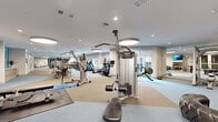 This expansive, single-floor fitness center offers a comprehensive workout environment thoughtfully designed to accommodate a wide range of exercise disciplines. The open layout is organized into distinct functional zones, allowing users to move seamlessly between cardio, strength, and functional training areas without crowding.

The cardio zone features a row of treadmills and elliptical machines positioned along floor-to-ceiling windows that invite abundant natural light and offer views to the outdoors, creating an energizing workout atmosphere. Adjacent to this area, a dedicated strength training section houses a full complement of Precor cable machines, multi-station weight systems, and barbell squat racks with bench press setups, all reflected in large wall-to-wall mirrors that enhance the sense of space.

A functional training area provides ample open floor space for stretching and bodyweight exercises, equipped with rowing machines, medicine balls, resistance bands, and stability equipment. A dumbbell rack with a full range of free weights completes the strength offerings.

The interior design is clean and contemporary, featuring neutral-toned rubber flooring, white ceilings with recessed lighting accented by distinctive round pendant fixtures, and accent walls in a calming blue tone. Wall-mounted flat-screen televisions are positioned throughout. Built-in shelving and a dedicated reception or check-in area near the entrance add to the facility's polished, professional character.
