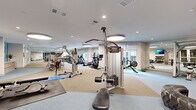 This expansive, single-floor fitness center offers a comprehensive workout environment thoughtfully designed to accommodate a wide range of exercise disciplines. The open layout is organized into distinct functional zones, allowing users to move seamlessly between cardio, strength, and functional training areas without crowding.

The cardio zone features a row of treadmills and elliptical machines positioned along floor-to-ceiling windows that invite abundant natural light and offer views to the outdoors, creating an energizing workout atmosphere. Adjacent to this area, a dedicated strength training section houses a full complement of Precor cable machines, multi-station weight systems, and barbell squat racks with bench press setups, all reflected in large wall-to-wall mirrors that enhance the sense of space.

A functional training area provides ample open floor space for stretching and bodyweight exercises, equipped with rowing machines, medicine balls, resistance bands, and stability equipment. A dumbbell rack with a full range of free weights completes the strength offerings.

The interior design is clean and contemporary, featuring neutral-toned rubber flooring, white ceilings with recessed lighting accented by distinctive round pendant fixtures, and accent walls in a calming blue tone. Wall-mounted flat-screen televisions are positioned throughout. Built-in shelving and a dedicated reception or check-in area near the entrance add to the facility's polished, professional character.