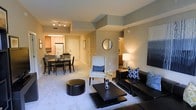 This well-appointed single-floor residence offers a seamlessly connected living experience through its open-concept layout. The living area serves as the heart of the home, featuring plush carpeted flooring, a sleek dark leather sofa accented with geometric throw pillows, a modern square coffee table, and a round accent chair. A circular mirror and contemporary wall art add decorative character to the neutral-toned walls, which are painted in a soft sage and warm beige palette.

The dining area flows naturally from the living space, accommodating a dark wood dining table with matching chairs — ideal for both everyday meals and entertaining. Just beyond, the kitchen is outfitted with light wood cabinetry, standard appliances including a refrigerator, and a functional layout that keeps everything within easy reach.

Track lighting in the kitchen and recessed ceiling lights throughout provide ample illumination, contributing to a bright and welcoming ambiance. A hallway leads to additional rooms, suggesting a thoughtful floor plan that balances open communal areas with private spaces. The overall aesthetic is clean, modern, and cohesive, making this a comfortable and visually appealing home.