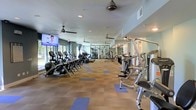 This fitness center offers a spacious, single-floor layout designed to accommodate a comprehensive workout experience. The room is generously proportioned, allowing ample space between equipment for comfortable movement throughout. Along one side, a row of cardio machines — including ellipticals, treadmills, and stair climbers — lines the wall beneath a mounted flat-screen television, making it easy to stay entertained during endurance training. On the opposite side, a selection of strength and cable machines provides options for resistance and weight training. Large windows along the far wall invite abundant natural light, creating an open and energizing atmosphere. Multiple ceiling fans are evenly distributed throughout the space, promoting airflow and comfort during intense workouts. The flooring consists of durable carpet tiles in a two-tone pattern of brown and blue, offering both cushioning and visual interest. Recessed lighting supplements the natural light, ensuring the space remains well-lit at all times. A mirrored section at the back of the room enhances the sense of depth and allows users to monitor their form. Overall, the fitness center presents a clean, functional, and well-maintained environment suited for a wide range of exercise routines.