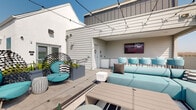 Located at 1575 Camino Altezza in Chula Vista, CA, this rooftop terrace is a well-appointed outdoor amenity space designed for both relaxation and social gatherings. The layout is divided into distinct functional zones, each thoughtfully furnished and connected by large-format tile flooring throughout.

The covered lounge zone anchors the space with an expansive sectional sofa in teal upholstery, paired with a low-profile coffee table and ottomans. A wall-mounted flat-screen television is positioned above a white subway-tiled bar counter, lined with matching teal bar stools. The covered overhang provides shade while maintaining an open connection to the outdoors.

An open-air lounge area features globe string lights overhead, a central fire pit surrounded by teal accent chairs and side tables, and striped area rugs adding visual definition. This zone offers unobstructed panoramic views of the hills and surrounding Chula Vista neighborhood, making it an ideal gathering spot at any time of day.

Additional seating is provided through sculptural lattice lounge chairs with teal cushions, positioned near raised planters with mature trees. The overall design palette — white and gray building exteriors, teal furnishings, and natural tones — creates a cohesive, modern aesthetic that complements the Southern California setting.