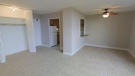 This single-floor residence offers a practical and comfortable layout designed for easy everyday living. The main living area is finished with soft wall-to-wall carpeting in a neutral tone, creating a warm and inviting atmosphere throughout. A ceiling fan overhead provides both comfort and a subtle design accent to the space.

The kitchen is thoughtfully positioned adjacent to the living area, separated by a partial wall that features a convenient breakfast bar — ideal for casual dining or additional counter space. The kitchen is equipped with wood-toned cabinetry, a white refrigerator, and a range, with tile flooring that contrasts neatly with the carpeted living space. The open pass-through design between the kitchen and living area enhances the sense of connectivity and allows for easy interaction between the two spaces.

A closet with bifold doors is visible near the entryway, offering convenient built-in storage. The clean white trim, neutral walls, and consistent color palette throughout contribute to a cohesive and understated aesthetic. The overall layout is efficiently organized, making the most of the available space while maintaining an open and uncluttered feel.