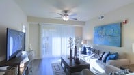 This single-floor space offers a comfortable and well-lit environment designed for modern living. The living area serves as the centerpiece, featuring a ceiling fan with an integrated light fixture that promotes year-round comfort and airflow. Large sliding glass doors dressed with vertical blinds span much of one wall, allowing an abundance of natural light to pour into the room and creating a bright, airy ambiance throughout the day. The hard-surface flooring adds a sleek, easy-to-maintain foundation that complements the neutral wall tones. A stylish area rug anchors the seating arrangement, which includes a plush sofa adorned with decorative throw pillows in coordinating blue and white tones. A dark-finished coffee table serves as a functional focal point at the center of the living space, accessorized with decorative vases and botanicals. A vibrant blue abstract artwork on the wall adds a pop of color and personality to the room. The overall aesthetic is clean, contemporary, and inviting, with thoughtful attention to lighting and décor that creates a relaxed yet polished atmosphere well-suited for both everyday living and entertaining.