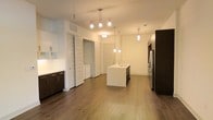This single-floor space showcases a thoughtfully designed open-concept layout that seamlessly connects the kitchen and living areas. The kitchen serves as a central focal point, featuring a white center island with an integrated sink, complemented by sleek dark-toned cabinetry that provides a striking contrast against the bright white walls and countertops. Stainless steel appliances add a polished, contemporary finish to the space.

Warm hardwood-style flooring runs throughout, lending continuity and a welcoming feel to the entire floor plan. Overhead, a modern linear pendant light fixture anchors the kitchen island, while additional hanging pendant lights illuminate the surrounding space with a clean, ambient glow. Recessed lighting further enhances the bright, airy atmosphere of the interior.

The open layout allows for a generous flow between functional zones, with multiple doorways visible in the background suggesting well-organized access to additional rooms. White paneled doors and neutral wall tones keep the aesthetic cohesive and versatile. Overall, the space presents a contemporary, move-in-ready environment with a clean design sensibility.