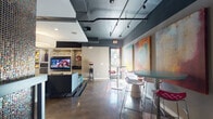 Located at 2505 Longview St in Austin, Texas, this single-floor communal amenity space offers a dynamic and stylish environment designed for social gatherings and relaxation. The interior showcases an industrial-modern aesthetic, highlighted by exposed dark-painted ductwork on the ceilings, polished concrete floors, and a bold color palette that blends deep charcoals, grays, and vibrant accent tones.

The main lounge area is anchored by a large built-in entertainment wall with a flat-screen television, custom cabinetry, and a plush sectional sofa arrangement beneath a statement drum pendant light. Adjacent to this is a casual dining zone featuring bar-height tables and colorful modern stools, positioned near a glass entry door that invites natural light into the space.

A standout architectural feature is a floor-to-ceiling column clad in iridescent mosaic tile, adding a striking visual focal point to the bar and high-top seating area. Large-scale abstract artwork adorns multiple walls throughout, contributing to the space's gallery-like energy.

A dedicated game room area includes a billiards table, lounge seating with eclectic upholstered ottomans, a computer workstation nook, and wall-mounted televisions — all set against bold, graphic wall murals. The space is illuminated by a combination of track lighting and recessed fixtures, creating a well-lit yet atmospheric environment suited for both casual and social use.