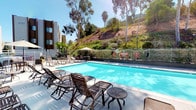Located at 6675 Jafte Ct in San Diego, this outdoor pool area offers a well-appointed recreational retreat set against a backdrop of natural hillside terrain and mature eucalyptus and palm trees. The centerpiece of the space is a generously sized swimming pool with clear, blue water, designed to accommodate both leisurely swims and relaxed sunbathing. The surrounding deck is finished in light-toned concrete, providing ample space for circulation and lounging. Numerous slatted chaise lounges and side tables are arranged throughout the deck, offering comfortable spots for sunbathing or unwinding poolside. Several large striped umbrellas provide shaded seating areas, ensuring comfort during San Diego's sun-filled days. The perimeter of the pool area is enclosed by a metal railing fence, with lush low-growing shrubs and colorful plantings softening the boundary and enhancing the overall aesthetic. The hillside beyond the fence, dotted with tall trees and natural vegetation, creates a scenic and private natural backdrop. The surrounding multi-story residential buildings with neutral-toned facades frame the space, contributing to its well-maintained and community-oriented character. The entire area benefits from abundant natural light and an open-air setting that captures the essence of Southern California outdoor living.