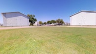 This single-story property is defined by its generous open outdoor grounds, offering a sense of space and openness that is immediately apparent. The exterior landscape is dominated by a broad, well-kept lawn that stretches across the property, providing an expansive green setting that contributes to the overall open and airy feel of the space.

Flanking the central green area are large, light-colored buildings with siding exteriors, which lend a clean and functional aesthetic to the property. Their substantial scale underscores the generous proportions of the surrounding grounds. Mature trees are scattered throughout and along the perimeter, offering natural shade and a softening contrast to the built structures nearby.

A paved driveway or roadway runs along the edge of the property, with additional structures and parked vehicles visible in the mid-distance, suggesting an active and accessible location within a broader community or commercial area. The surroundings indicate a flat, open landscape characteristic of a plains or rural setting.

The property benefits from abundant natural light, with clear skies that enhance the brightness and openness of the outdoor environment. The combination of manicured lawn, mature trees, and well-maintained neighboring structures creates a tidy, functional, and welcoming exterior setting suited for a variety of uses.