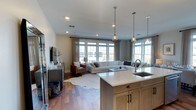 Located at The Vanderbilt on Corporate Drive in Westbury, New York, this single-floor residence offers a thoughtfully designed open-concept layout that seamlessly connects the kitchen, dining, and living areas into one cohesive living space. The kitchen is anchored by a substantial island with a white quartz countertop and an undermount sink, complemented by warm natural wood cabinetry with matte black hardware. Pendant lighting with glass shades hangs above the island, adding both functional illumination and a refined decorative touch. Recessed lighting throughout reinforces the bright, airy atmosphere. The living area is generously sized, accommodating a large sectional sofa and accent seating, all grounded by a cowhide-style area rug over rich hardwood flooring that extends across the entire space. A wall of large, multi-pane windows along the far side floods the interior with natural light and provides views of the surrounding area. Sheer curtains frame the windows, softening the light while maintaining an open feel. The neutral color palette of whites, warm grays, and natural wood tones contributes to a clean, modern aesthetic, making this a welcoming and well-appointed residence.