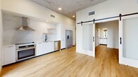 Located at 2281 N Lois Ave in Tampa, FL, this thoughtfully designed unit blends modern industrial aesthetics with comfortable living. The open-concept main area features warm hardwood-style flooring and a striking exposed concrete ceiling with recessed lighting and a ceiling fan, creating an airy, loft-inspired atmosphere. Floor-to-ceiling sliding glass doors flood the living space with natural light and open directly onto a private balcony, where black metal railings frame views of lush tropical landscaping, mature palms, and manicured garden pathways. The kitchen is outfitted with sleek white cabinetry, marble-style backsplash, stainless steel appliances including a French door refrigerator and dishwasher, and a deep basin sink — all finished with a clean, contemporary aesthetic. A frosted glass sliding barn door adds a stylish functional detail, separating the living areas from the hallways with understated elegance. The bathroom features a soaking tub with shower, smooth large-format wall surround, and matte black fixtures that complement the unit's modern design language. Light-filled hallways with recessed lighting connect all areas seamlessly. This well-appointed space offers a cohesive blend of industrial design, natural light, and Florida-inspired outdoor living.