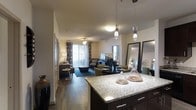 This single-floor residence showcases a thoughtfully designed open-concept layout that seamlessly connects the kitchen, dining, and living areas into one cohesive living space. The kitchen is anchored by a substantial granite-topped island with dark cabinetry beneath, providing generous prep and entertaining space. Elegant pendant lighting hangs above the island, adding a refined touch to the culinary space. The dining area flows naturally from the kitchen, offering a dedicated space for gatherings, while decorative mirrors enhance the sense of openness. Rich dark hardwood flooring extends throughout, grounding the interior with warmth and continuity. The living room is generously proportioned, bathed in natural light streaming through floor-to-ceiling sliding glass doors that lead to an outdoor patio or balcony. Vertical blinds offer privacy and light control while maintaining the room's airy feel. Neutral wall tones throughout provide a versatile backdrop for a variety of interior styles. The overall ambiance strikes a balance between contemporary sophistication and comfortable everyday living, making this a well-appointed and functional single-level home.