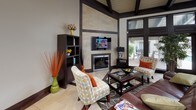 This single-floor space showcases a thoughtfully designed interior that blends contemporary style with warm, inviting finishes throughout. The living area is a standout, boasting dramatically high ceilings with exposed dark wood beams and a striking floor-to-ceiling tile surround framing a sleek fireplace. A wall-mounted flat-screen television anchors the space, while large dark-framed glass doors and transom windows flood the room with natural light and offer views of an outdoor patio area. The tile flooring features an elegant inlaid dark wood border pattern that adds visual definition and continuity across the space. The kitchen is well-equipped and functional, featuring rich dark wood cabinetry with brushed nickel hardware, granite countertops, stainless steel appliances including a full-size refrigerator and built-in microwave, and a breakfast bar that provides additional prep and seating space. The soaring ceilings carry through into the kitchen, enhancing the open, airy feel. Dark wood trim, baseboards, and door frames are consistent throughout, tying together the design language of the space and creating a cohesive, refined aesthetic. Natural light, generous ceiling height, and high-quality finishes combine to give this space a warm yet sophisticated character.