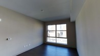 This single-floor space presents a clean, modern aesthetic defined by its understated yet thoughtfully considered design elements. The room features rich, dark hardwood flooring that adds warmth and contrast against the smooth, light-toned walls and white ceiling. A large, multi-pane window serves as a striking focal point, flooding the interior with natural light while framing an expansive view of the surrounding urban cityscape — including neighboring buildings, streets, and the city skyline beyond. The elevated vantage point suggested by the window view reinforces the space's position within a larger multi-story building. The walls are finished in a neutral, warm beige tone that complements the dark flooring and contributes to a calm, versatile ambiance. The ceiling is clean and unadorned, maintaining a sense of openness throughout the room. Overall, the space offers a bright, contemporary environment well-suited for a variety of uses, with its city-facing window and quality finishes standing out as defining characteristics.