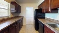 This single-story space presents a clean, functional layout with a neutral color palette throughout. The living areas are finished with wall-to-wall beige carpeting, creating a cohesive and comfortable feel across multiple rooms. Natural light enters through blinded windows, brightening the open, simply designed interiors. A ceiling fan with integrated lighting fixture is installed in one of the carpeted rooms, adding both ventilation and ambient light. An exterior door with a glass panel provides additional light to one end of the living space, with a tiled entry transition at the threshold.

The kitchen follows a galley-style configuration, featuring rich dark wood cabinetry with brushed hardware on both upper and lower cabinets. Granite-style countertops run the length of the kitchen, offering generous workspace. The flooring transitions to neutral ceramic tile, and black appliances—including a refrigerator and dishwasher—are present. A window above the counter brings in natural light. A gas range is also included.

The bathroom includes a full tub-and-shower combination with white square ceramic tile surround, a grab bar, standard fixtures, and a wall-mounted mirror above the vanity area. Overall, the space maintains a practical, move-in-ready presentation.