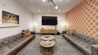 Located at 221 31st Ave N in Nashville, TN, this single-floor amenity space offers a thoughtfully designed collection of social and leisure areas, each distinguished by its own personality and purpose. The layout flows seamlessly between zones, unified by patterned carpeting, recessed lighting, and a cohesive palette of warm neutrals accented by bold pops of color.

The media lounge serves as a central gathering point, featuring two tufted gray sofas flanking a large wall-mounted flat-screen television, complemented by gold-accented round coffee tables and a wooden media console. A vivid orange-and-red patterned accent wall adds a retro-inspired energy to the space.

A dedicated game room provides a standout experience, centered around a custom poker table with built-in chip holders, surrounded by upholstered wingback chairs. A striking geometric wallcovering in gold, gray, and black anchors the room, while framed playing card artwork adds a playful, thematic touch.

A relaxed seating nook features two white lounge chairs, a side table with a decorative clock, and a bold yellow framed artwork, set against a vibrant floral-patterned accent wall. A semi-private workspace with a solid wood desk and blue leather chairs opens to an adjacent dining or meeting area with dark upholstered seating. Dark-framed glass double doors with transom panels define the entryway, completing this well-appointed amenity suite.