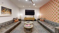 Located at 221 31st Ave N in Nashville, TN, this single-floor amenity space offers a thoughtfully designed collection of social and leisure areas, each distinguished by its own personality and purpose. The layout flows seamlessly between zones, unified by patterned carpeting, recessed lighting, and a cohesive palette of warm neutrals accented by bold pops of color.

The media lounge serves as a central gathering point, featuring two tufted gray sofas flanking a large wall-mounted flat-screen television, complemented by gold-accented round coffee tables and a wooden media console. A vivid orange-and-red patterned accent wall adds a retro-inspired energy to the space.

A dedicated game room provides a standout experience, centered around a custom poker table with built-in chip holders, surrounded by upholstered wingback chairs. A striking geometric wallcovering in gold, gray, and black anchors the room, while framed playing card artwork adds a playful, thematic touch.

A relaxed seating nook features two white lounge chairs, a side table with a decorative clock, and a bold yellow framed artwork, set against a vibrant floral-patterned accent wall. A semi-private workspace with a solid wood desk and blue leather chairs opens to an adjacent dining or meeting area with dark upholstered seating. Dark-framed glass double doors with transom panels define the entryway, completing this well-appointed amenity suite.