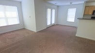 This single-floor space features an open and airy living area finished with neutral-toned wall-to-wall carpeting and light gray-beige walls that create a clean, understated aesthetic. The room is generously proportioned, offering a versatile layout suitable for a variety of furniture arrangements. A standout feature is the set of French doors, which introduce natural light while providing access to the outdoors. An additional window on the adjacent wall further brightens the interior, contributing to the overall sense of openness. The ceiling is standard height and includes a centrally placed light fixture, along with a visible air vent for climate control. The simple, unadorned walls provide a blank canvas, making the space highly adaptable to different interior design styles. The soft carpeting throughout adds warmth and comfort underfoot, complementing the light and neutral color palette. Overall, the space presents a functional and welcoming environment with practical amenities and a straightforward layout well-suited for everyday living.