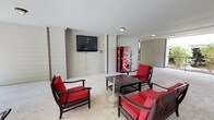 This single-story covered communal space serves as a shared gathering area, thoughtfully designed to blend indoor comfort with outdoor accessibility. The space is defined by horizontal lap siding in neutral tones accented by dark brown structural columns, giving it a clean, residential character throughout.

The interior is organized into distinct seating zones. One area features dark wood-framed furniture with bold red cushions arranged around a low coffee table, creating an inviting social setting. A wall-mounted flat-screen television is positioned nearby, making it suitable for casual viewing. A second grouping of wicker chairs with colorful cushions provides an alternative relaxation spot. Vending machines are conveniently positioned along the wall for resident use.

Open on one side, the space transitions toward a railed terrace overlooking lush greenery and mature trees, allowing natural light and fresh air to flow freely throughout. Flush-mount ceiling fixtures provide supplemental lighting for evening use.

A separate covered patio area features distinctive arched openings that frame views of the manicured lawn and surrounding buildings. This patio includes a round metal outdoor dining table and chairs, ideal for al fresco gatherings. The combination of open-air accessibility, comfortable furnishings, and green outdoor views makes this a versatile and welcoming communal retreat.