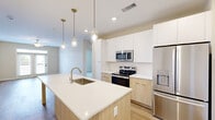 This thoughtfully designed 2-bedroom, 3-bathroom residence at 2418 Main St in Rocky Hill, Connecticut offers a clean, modern aesthetic throughout its single-floor layout. The open-concept kitchen serves as the heart of the home, featuring a large waterfall-edge island with an undermount sink, crisp white upper cabinetry paired with warm natural wood lower cabinets, stainless steel appliances including a French door refrigerator, electric range, and built-in microwave, all illuminated by a trio of elegant glass pendant lights. Light hardwood-style flooring flows seamlessly through the kitchen and living areas, complementing the neutral color palette. The spacious living area is bathed in natural light through French doors that open to a balcony, with a ceiling fan adding comfort overhead. Both bedrooms are carpeted for warmth and each is equipped with a ceiling fan and window with exterior views. The generously sized primary bedroom includes an en-suite bathroom with a wood-toned vanity, white quartz countertop, stylish gold sconce lighting, and access to a walk-in closet with wire shelving systems. Two additional closets provide ample storage throughout. Clean white panel doors with matte black hardware add a refined, contemporary touch to every room.