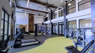 Located at 1040 Wake Towne Dr in Raleigh, NC, this contemporary fitness center delivers a well-appointed workout environment designed to accommodate a wide range of exercise needs. The space makes an immediate impression with its soaring double-height ceilings, exposed ductwork, and industrial-style large ceiling fans that lend the facility a modern, open aesthetic. Warm wood accent panels contrast thoughtfully against crisp white walls and dark structural columns, creating a visually dynamic atmosphere that feels both energizing and welcoming. Large windows and glass doors along the perimeter invite abundant natural light throughout the space, reinforcing the sense of openness. The fitness floor is thoughtfully zoned, featuring a bright yellow rubberized activity surface that delineates dedicated training areas. Equipment includes multiple treadmills, cable and strength machines, a squat rack, and a variety of free weights, ensuring comprehensive options for both cardio and resistance training. Mirrored wall sections enhance the sense of space and support form-checking during workouts. The overall layout promotes ease of movement between stations, making the facility functional for individual workouts as well as group training sessions. This is a well-designed, full-featured fitness facility built to serve residents and users seeking a high-quality exercise experience.