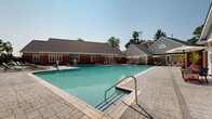 Located at 8130 Prescott Dr in Vienna, Virginia, this community pool facility offers a well-appointed outdoor aquatic and recreation space spread across two floors. The centerpiece is a generously sized outdoor swimming pool with clearly marked depth gradations ranging from a shallow end of 3.5 feet to a deep end of 8.5 feet, making it suitable for a wide range of swimmers. The pool features stainless steel entry rails and is finished with classic tiled accents along the waterline.

Surrounding the pool is an expansive stamped concrete sundeck, thoughtfully lined with rows of modern lounge chairs that provide ample space for sunbathing and relaxation. Shaded seating areas with patio umbrellas and outdoor furniture are positioned around the perimeter, offering comfortable alternatives for those seeking respite from the sun.

The adjacent clubhouse structure is a well-maintained two-floor building combining brick and shingle siding exteriors, accented with large multi-pane windows and pitched rooflines. Restroom and utility access facilities are conveniently located along the poolside exterior of the building. Mature trees frame the background of the property, providing natural shade and a lush green backdrop. The overall setting is clean, well-maintained, and designed to serve as an inviting community gathering space throughout the warmer months.