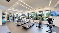 Located at 1340 S Dixie Hwy in Coral Gables, Florida, this single-floor fitness center is a well-appointed facility designed to support a comprehensive workout experience. The space is defined by its clean, contemporary aesthetic, featuring crisp white walls, recessed linear LED lighting along the ceiling, and durable rubber flooring throughout the main exercise area. Large picture windows line the perimeter, flooding the interior with natural light and offering views of the surrounding tropical landscaping, creating an energizing and open atmosphere. The cardio zone is equipped with multiple treadmills, elliptical trainers, stationary bikes, and a rowing machine, each outfitted with personal display screens. The strength training area features a multi-functional cable machine and a full wall of mirrors that amplify the sense of space. Wall-mounted flat-screen TVs provide entertainment throughout. The entry corridor is finished with warm wood-look flooring and includes a built-in hand sanitizer dispenser and a stainless steel water fountain station, reflecting a strong emphasis on hygiene and convenience. Separate restroom facilities are accessible directly from the entry hallway. The facility entrance features a glass-paneled door branded with the property logo, offering a glimpse of the outdoor pool area beyond.
