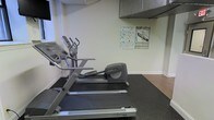 This single-level fitness room is designed to provide a functional workout space with a focused selection of cardio equipment. The room features a Life Fitness treadmill and an elliptical trainer positioned on durable rubber flooring, which protects the subfloor and provides stability during exercise. The walls are finished in a neutral, light tone, contributing to a clean and straightforward environment. Informational fitness charts, including a heart rate target reference guide and an exercise diagram, are mounted on the wall, offering users helpful guidance during their workouts. Overhead, a metal ventilation unit is visible, indicating climate control provisions to maintain comfort during physical activity. The layout is efficient, making practical use of the available space to accommodate the equipment without overcrowding. The overall ambiance is utilitarian and purpose-driven, suited for residents or occupants seeking convenient access to basic cardio exercise equipment without the need to visit an off-site facility.