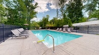 Located at 9 Covered Bridge Rd in Newtown, Connecticut, this outdoor pool area offers a well-appointed and tranquil recreational space surrounded by lush, mature trees that provide a natural sense of privacy and seclusion. The centerpiece is a generously sized rectangular swimming pool with clear, blue-tinted water and tiled accent detailing along the floor. The pool is accessible via built-in handrails and entry steps, making it welcoming for a range of users. Encircling the pool is a spacious stone-paved deck outfitted with a full row of reclining lounge chairs, ideal for relaxing in the sun. Umbrella-shaded seating areas are positioned throughout the deck, offering comfortable spots for outdoor dining and leisure. A secondary seating zone with a table, chairs, and a large canopy umbrella provides additional shade and a more intimate gathering area. The entire pool area is enclosed by a sturdy black metal fence, ensuring safety and security. An adjacent single-story pool house structure with a neutral facade and pitched roof provides utility support. The surrounding canopy of tall deciduous trees creates a serene, park-like atmosphere beneath open skies, making this outdoor space a refreshing and functional amenity.