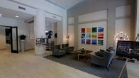 This single-floor lobby space makes an immediate impact with its soaring ceilings and open, airy layout. A prominent textured column anchors the room, adding architectural interest and a tactile contrast to the smooth surrounding walls. The space is thoughtfully designed with distinct seating areas, each furnished with plush, charcoal-toned upholstered chairs and sofas arranged around a sleek wooden coffee table adorned with a simple floral centerpiece.

A bold grid of nine colorful framed photographs dominates one wall, serving as a striking focal point that introduces vibrant hues—yellow, blue, red, and purple—against the otherwise neutral palette of soft grays and warm whites. A sculptural Sputnik-style pendant light adds a retro-modern touch, complementing the contemporary aesthetic throughout.

The flooring transitions from light hardwood to a patterned area rug beneath the seating arrangement, defining the lounge zone within the open plan. A display easel near the entrance provides informational or directional content for visitors. Overall, the space balances sophisticated design with functional comfort, projecting a professional yet inviting ambiance.