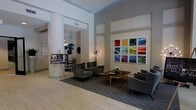 This single-floor lobby space makes an immediate impact with its soaring ceilings and open, airy layout. A prominent textured column anchors the room, adding architectural interest and a tactile contrast to the smooth surrounding walls. The space is thoughtfully designed with distinct seating areas, each furnished with plush, charcoal-toned upholstered chairs and sofas arranged around a sleek wooden coffee table adorned with a simple floral centerpiece.

A bold grid of nine colorful framed photographs dominates one wall, serving as a striking focal point that introduces vibrant hues—yellow, blue, red, and purple—against the otherwise neutral palette of soft grays and warm whites. A sculptural Sputnik-style pendant light adds a retro-modern touch, complementing the contemporary aesthetic throughout.

The flooring transitions from light hardwood to a patterned area rug beneath the seating arrangement, defining the lounge zone within the open plan. A display easel near the entrance provides informational or directional content for visitors. Overall, the space balances sophisticated design with functional comfort, projecting a professional yet inviting ambiance.