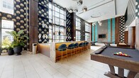 Located at 715 US Highway 46, this sophisticated amenity lounge spans a single, expansive floor and is designed to impress with its bold, contemporary aesthetic. Soaring double-height ceilings amplify the sense of openness, while floor-to-ceiling windows flood the space with natural light and provide views of the outdoor terrace area beyond.

The interior is defined by striking design details throughout. A dramatic feature wall clad in teal-glazed brick tile serves as a focal point, flanked by illuminated vertical accents and a mounted flat-screen display, with plush channeled sofas arranged for comfortable lounge seating. A signature botanical wallpaper in navy and gold repeats across multiple walls, lending cohesion and a luxurious character to the space.

A central wet bar with a stone-topped counter and glowing amber base is lined with velvet-upholstered barstools in deep blue with brass legs, creating an inviting social hub. Adjacent built-in shelving with warm wood finishes displays decorative ceramics and glassware. A separate high-top seating area with a wall-mounted TV offers an additional gathering spot.

The recreation zone features a full billiards table and two ping-pong tables set atop a graphic black-and-white patterned rug. Mirrored globe pendant lights and recessed ceiling fixtures complete the refined yet playful ambiance throughout.