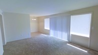 This single-floor space offers a clean, open layout filled with natural light. The main living area is generously sized, featuring wall-to-wall carpeting in a neutral tone that lends a warm and comfortable feel underfoot. Large vertical blinds frame a sliding glass door, allowing ample daylight to pour into the room while offering easy access to the outdoors. An additional window further enhances the brightness of the space.

Adjacent to the living area, a defined dining space is elegantly appointed with a chandelier, adding a refined touch to the otherwise understated interior. The walls are finished in a soft, neutral palette that complements the light carpeting and creates a cohesive, airy ambiance throughout.

The open flow between the living and dining areas makes the space feel expansive and versatile, well-suited for both everyday living and entertaining. The minimal, unfurnished state of the space allows its architectural layout and natural light to take center stage, giving prospective occupants a clear canvas to personalize as they see fit.