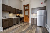 Located at 101 E Fifth Ave in Knoxville, Tennessee, this single-floor unit showcases a thoughtful blend of historic character and contemporary finishes. Soaring ceilings throughout create an expansive, airy atmosphere, while warm hardwood-style flooring flows seamlessly from room to room. The main living area is highlighted by an ornate coffered ceiling with decorative molding, a pendant light fixture, and tall windows that invite generous amounts of natural light. Neutral gray tones on the walls complement the light flooring for a clean, modern aesthetic. The kitchen is well-appointed with stainless steel appliances—including a range, over-the-range microwave, dishwasher, and refrigerator—paired with rich wood cabinetry and light countertops. A charming original wood-framed door with frosted glass transoms adds a distinctive historic touch to the kitchen entry. The bedroom features the same soaring ceilings, a tall window, and a classic paneled door. The bathroom includes a single vanity with ample cabinet storage, a large mirror, and a shower/tub combination, with a high window providing natural light. Additional storage is available through a walk-in closet. Throughout the space, bold crown molding and oversized baseboards reinforce the building's historic architectural roots.