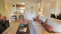 This single-floor residence offers a thoughtfully designed open-concept layout that promotes a natural flow between the living and kitchen areas. The living room is warm and inviting, featuring neutral-toned carpeting that adds softness underfoot, while crown molding along the ceiling lends a refined architectural touch. A comfortable seating area anchors the room, complemented by warm accent lighting from a stylish floor lamp. Decorative circular mirrors adorn the wall, adding visual interest and depth to the space.

The kitchen is accessible via a pass-through counter with bar seating, creating a casual and sociable dining option. The kitchen features white cabinetry and overhead lighting, with the pass-through bar area accented by a rich terracotta-toned wall that adds a warm, vibrant contrast to the otherwise neutral palette.

Throughout the space, the color scheme blends creamy whites, warm beiges, and earthy terracotta accents, creating a cohesive and welcoming ambiance. The open sightlines between rooms enhance the sense of spaciousness, making the layout feel airy and connected. Overall, this residence balances functional design with comfortable, homey aesthetics, making it an ideal space for both relaxing and entertaining.