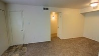 This single-floor residential space offers a straightforward and functional layout designed for comfortable everyday living. The interior is finished with neutral, light-toned walls throughout, creating a clean and airy atmosphere that lends itself well to a variety of décor styles. Wall-to-wall carpeting in a speckled, earthy tone runs across the main living areas, adding warmth and a soft underfoot feel to the space. A ceiling-mounted HVAC vent is integrated into the main room, ensuring efficient climate control. The rooms connect through open doorways, allowing for a smooth and natural flow between spaces. A wall-mounted sconce light fixture in the hallway area provides warm, ambient lighting, lending a welcoming quality to the transition between rooms. Standard white interior doors complement the clean aesthetic of the space. Electrical outlets and light switches are conveniently positioned throughout the walls. The overall design is simple and practical, with a move-in-ready feel suited to those looking for an uncluttered, low-maintenance living environment. The single-floor layout ensures ease of accessibility and navigation throughout the home.