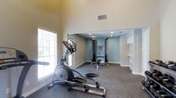 This single-floor fitness space is thoughtfully designed to support a complete workout routine in a comfortable, dedicated environment. The room features impressively high ceilings that lend an open, airy feel, while large windows fitted with blinds allow an abundance of natural light to fill the space throughout the day. The floor is covered in durable, low-pile carpet, providing a practical surface suited for exercise equipment and movement. The layout is divided into distinct functional zones: a cardio area near the windows houses an elliptical trainer, while a recessed section with recessed ceiling lighting serves as the strength training zone, equipped with a multi-function cable machine and a weight bench. The accent wall in the strength training area is painted in a soft blue tone, adding a visual distinction between the zones and contributing to a calm, motivating atmosphere. Built-in shelving and cabinetry on one wall offer convenient storage for accessories and equipment. Ventilation is supported by ceiling vents, ensuring comfort during extended workouts. Overall, the space combines practical functionality with a clean, organized aesthetic, making it a well-rounded fitness environment.