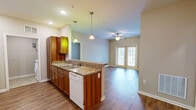 Located in Snellville, GA, this well-appointed 2-bedroom, 1-bathroom single-floor unit offers a thoughtfully designed layout across a comfortable footprint. The open-concept kitchen and living area serves as the heart of the home, featuring warm wood-toned cabinetry, granite-style countertops with a breakfast bar peninsula, pendant lighting, and hardwood-style flooring that flows throughout. The kitchen is fully equipped with a full-size refrigerator, electric range, microwave, and dishwasher, with an in-unit washer connection nearby for added convenience. French doors in the living area invite natural light and provide outdoor access. The spacious primary bedroom is outfitted with plush carpeting, a ceiling fan, double-door closet, and access to a generously sized walk-in closet with built-in wire shelving. A second bedroom, also carpeted and ceiling fan-equipped, offers a bright window with outdoor views. The shared bathroom features rich wood-toned cabinetry, granite countertops, a large vanity with ample storage, and tile flooring. A dedicated utility room completes the layout, enhancing overall functionality. Neutral wall tones and consistent finishes throughout lend the space a cohesive, move-in-ready appeal.