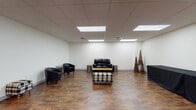 This single-floor space offers a well-designed, open-concept layout that blends functionality with a welcoming atmosphere. The most striking architectural feature is a large, floor-to-ceiling glass block wall near the main entrance, which diffuses natural light throughout the interior while maintaining privacy. Double wood-framed entry doors with narrow glass panels provide a clean, professional entryway, complemented by a dark floor mat.

The flooring throughout is a rich, dark wood-look surface that adds warmth and visual depth to the space. Overhead, a suspended tile ceiling is fitted with recessed rectangular LED light panels, ensuring consistent and even illumination across the entire area.

The main gathering area is furnished with a black leather loveseat and matching accent chairs arranged around a rustic industrial-style coffee table with a lower shelf. A plaid upholstered ottoman adds a touch of casual comfort to the seating arrangement. Cylindrical metallic side tables and decorative woven cone sculptures contribute to the space's eclectic yet cohesive aesthetic.

A secondary open area features a long table draped in a black cloth, ideal for catering, display, or registration use. The clean, neutral white walls throughout provide a versatile backdrop, making this space well-suited for a variety of events, meetings, or community gatherings.