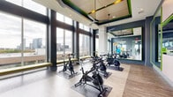 Located at Post Oak Park in the heart of Houston, TX, this dedicated exercise room is a well-appointed spin studio designed for focused, high-energy workouts. The space features soaring ceilings with a striking coffered detail accented in bold green, lending a dynamic, modern aesthetic throughout.

Floor-to-ceiling windows line an entire wall, flooding the room with natural light and offering panoramic views of the Houston skyline — an inspiring backdrop for every workout. Light wood-look flooring runs throughout, complementing the cool gray walls and vibrant green accent trim that define the room's contemporary design language.

Multiple stationary spin bikes are arranged across the studio floor, each positioned on individual floor mats for stability. Large wall-mounted mirrors with green-framed accents visually expand the space and allow users to monitor their form during exercise. A flat-screen TV is centrally mounted, ideal for guided cycling sessions or entertainment. Ceiling fans provide additional airflow throughout the room.

Built-in bench seating upholstered in chartreuse lines the window wall, offering a convenient spot for stretching or transitioning between exercises. Stability balls and additional fitness accessories are also available within the space. The overall ambiance is energetic yet polished, combining functional fitness design with an elevated, boutique studio atmosphere.