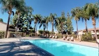 This outdoor community amenity area centers around a generously sized swimming pool with clear, blue-tinted water, surrounded by a wide concrete sundeck. The deck is well-equipped with multiple lounge chairs and seating arrangements positioned to take full advantage of the open sky and abundant Southern California sunshine.

Tall palm trees and mature evergreens line the perimeter of the pool area, creating a tropical, resort-like atmosphere while also providing natural shade and privacy. The pool enclosure is secured by a metal fence, enhancing both safety and exclusivity for residents.

Safety is clearly prioritized in this space, with posted pool rules, surveillance notices, and an emergency shutoff panel mounted on the textured stucco wall near the pool entrance.

Adjacent to the pool area is a private patio garden space featuring raised brick planters filled with lush, varied greenery, including ornamental shrubs and tropical plants. This quieter nook offers a more intimate outdoor retreat with seating for relaxation.

The surrounding residential buildings, finished in warm earth tones with pitched rooflines, frame the community grounds and contribute to the cohesive, well-maintained character of the overall space. The consistently sunny environment enhances the inviting, open-air ambiance throughout.